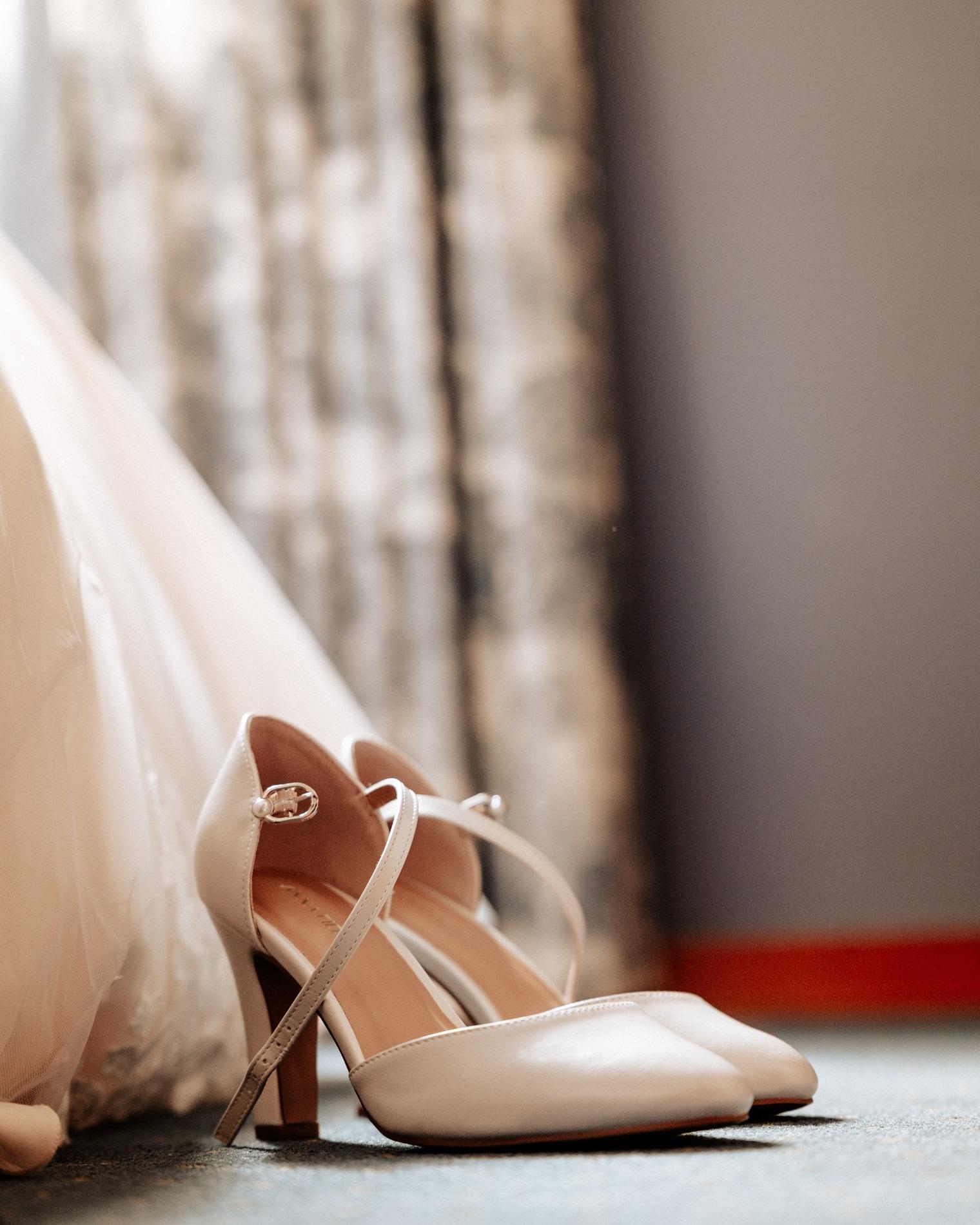 Cremefarbene Brautschuhe neben dem Saum des Brautkleids beim Getting Ready